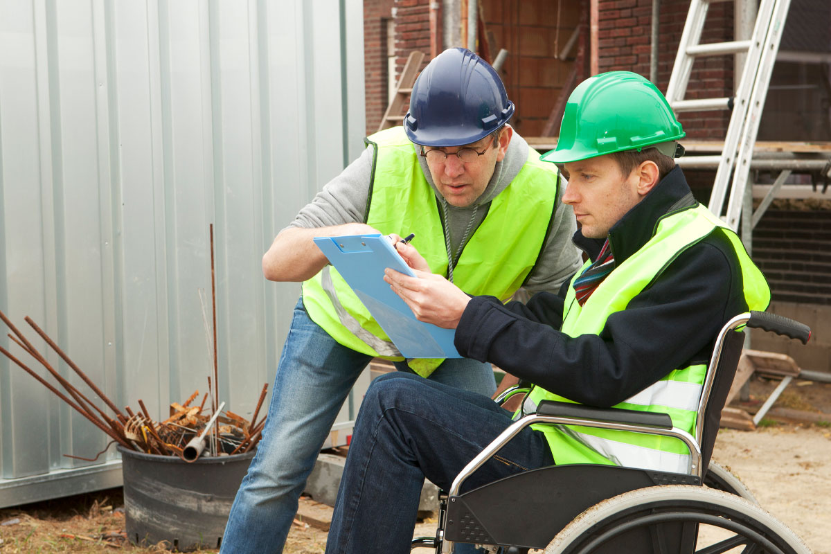 Auf einer Baustelle planen zwei Männer in Warnwesten einer im Rollstuhl die Arbeiten.