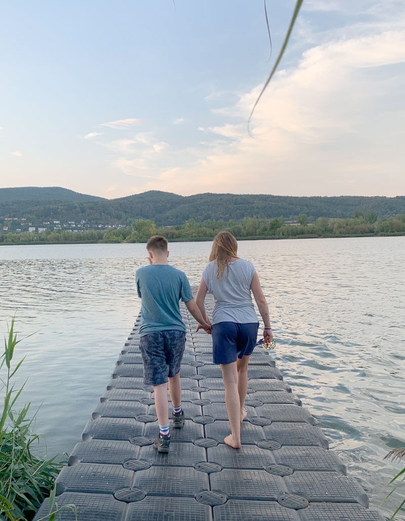 Ein Junge und eine Frau laufen Hand in Hand auf einem Steg am See. 